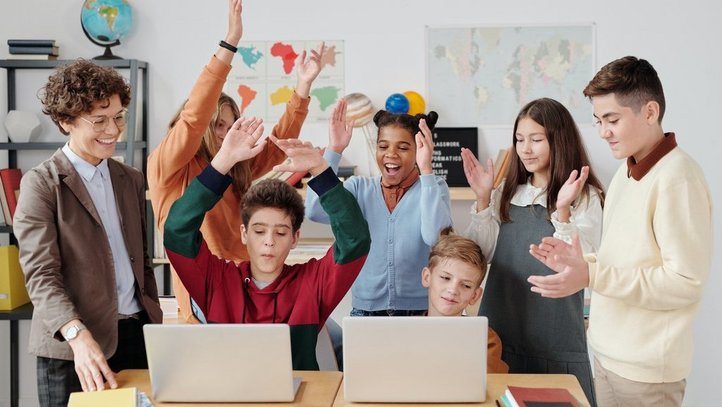 Schülerinnen und Schüler in einer Klasse mit ihrer Lehrerin, zwei Schüler sitzen vor einem Laptop und die Klasse freut sich.